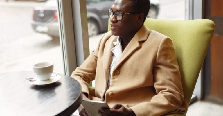 Serious African American male in trendy formal suit and eyeglasses sitting on cozy chair in cafe with cup of coffee and browsing tablet