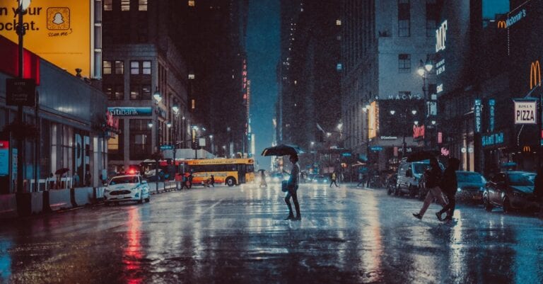 A rainy evening in New York City with people crossing a wet street under umbrellas.