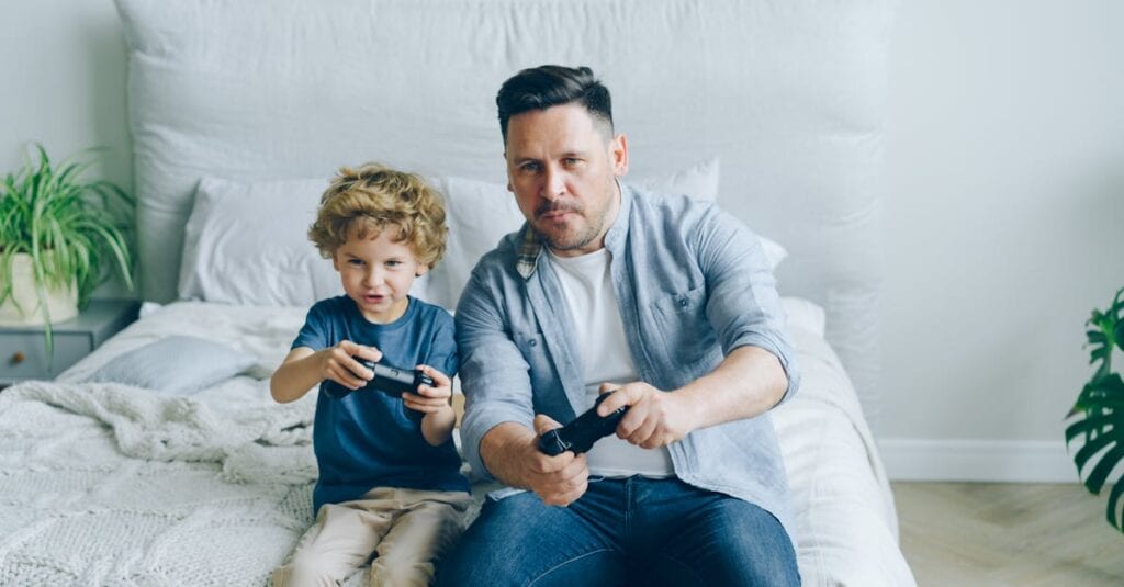 Father and son joyfully playing video games while sitting on a bed, capturing a moment of family fun.