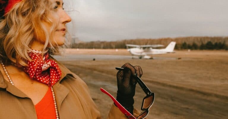 A fashionable woman with a red scarf and sunglasses standing near a small airplane.