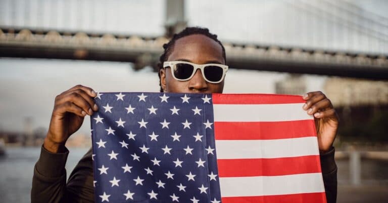 Car Service Blogs | Articles | News | Tips & Tricks | Video | FAQ | Information | Infograph. 40 Man holding an American flag in front of a bridge in New York City, showcasing patriotism.