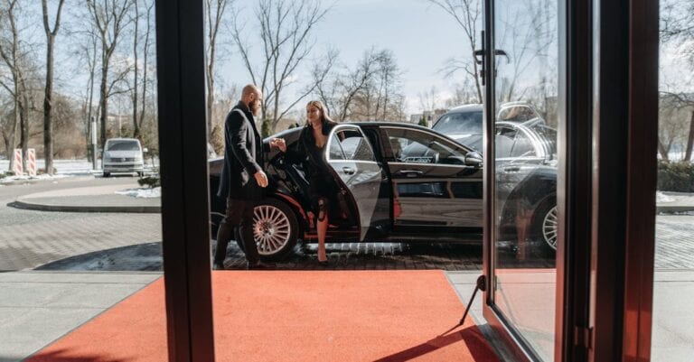A couple stepping out of a luxury car onto a red carpet in an upscale outdoor setting.