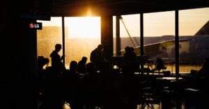 Travelers sitting in an airport terminal at sunset, silhouetted against the backdrop of an airplane.