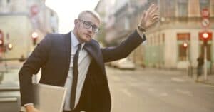 A confident businessman in a suit hails a taxi on a bustling city street, holding a laptop.