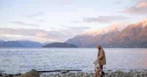 Side view of mother and little kid in casual warm outwear walking together along calm water on stony shore and holding hands