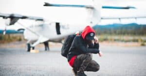 Full length unrecognizable male backpacker wearing casual outfit hunkering down against blurred modern light plane and touching cap peak before flight