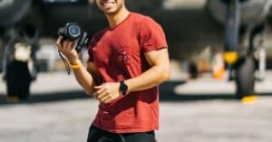 A cheerful photographer stands in front of a vintage aircraft, capturing moments on a sunny day.
