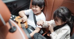 From above smiling ethnic boy and girl in casual outfits sitting fastened in passenger seats with ukulele during road trip together