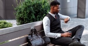 Young professional sitting on a bench, checking his wristwatch with coffee in hand.
