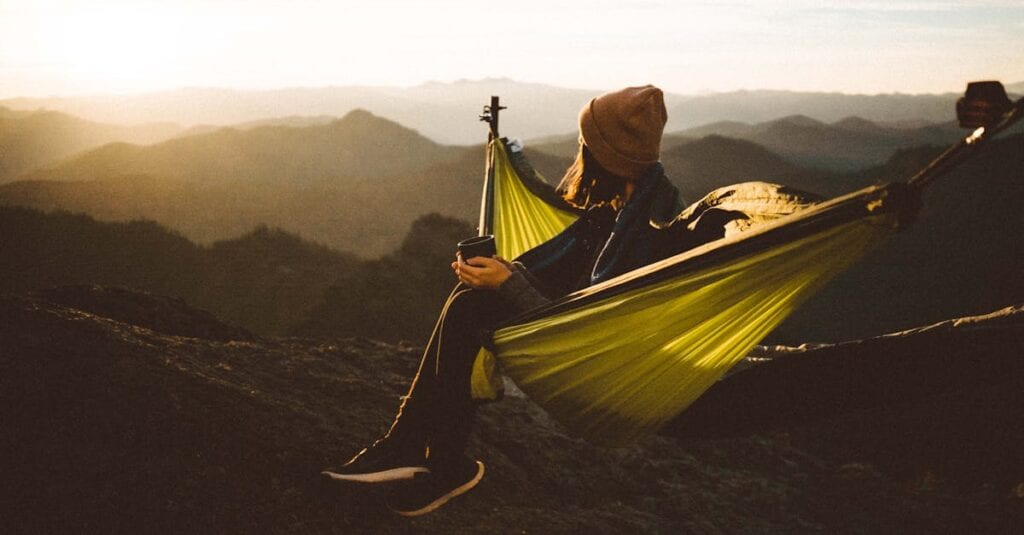 Woman relaxing in a hammock with a hot drink amidst majestic mountain scenery at sunset.