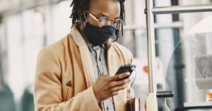 Stylish man with dreadlocks wearing a face mask on public transportation, checking his phone. Urban lifestyle context.