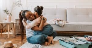 A mother and daughter share a loving hug while preparing for a trip with an open suitcase at home.
