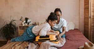 A mother and daughter pack a yellow suitcase in a cozy bedroom setting.