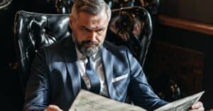 Elderly businessman in a suit reading a newspaper while sitting in a leather chair indoors.