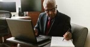 Elderly businessman in a suit writing notes while using a laptop in a modern office setting.