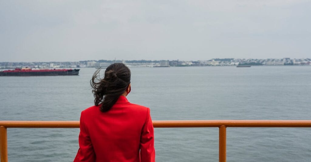 Woman in a red coat looking at the ocean from a ferry observation deck.