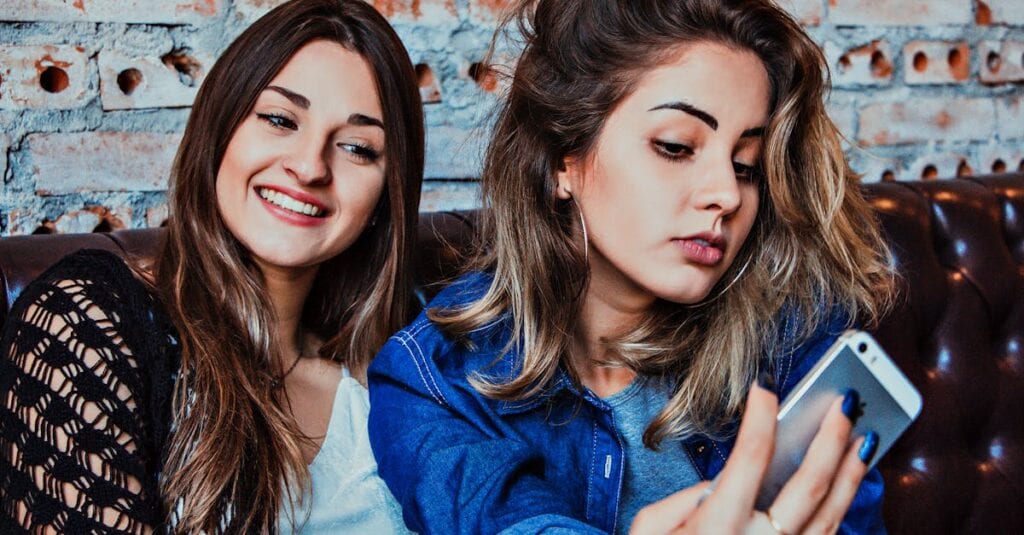 Two young women capturing a selfie while sitting in a casual indoor setting, smiling and relaxed.