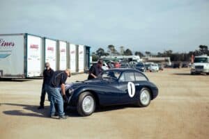 Three mechanics examining a vintage race car outdoors in Carmel-by-the-Sea, CA.
