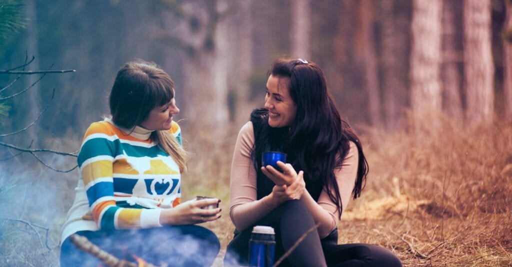 Two women enjoy coffee by a campfire in a forest, embracing the warmth and togetherness of nature.