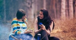 Two women enjoy coffee by a campfire in a forest, embracing the warmth and togetherness of nature.