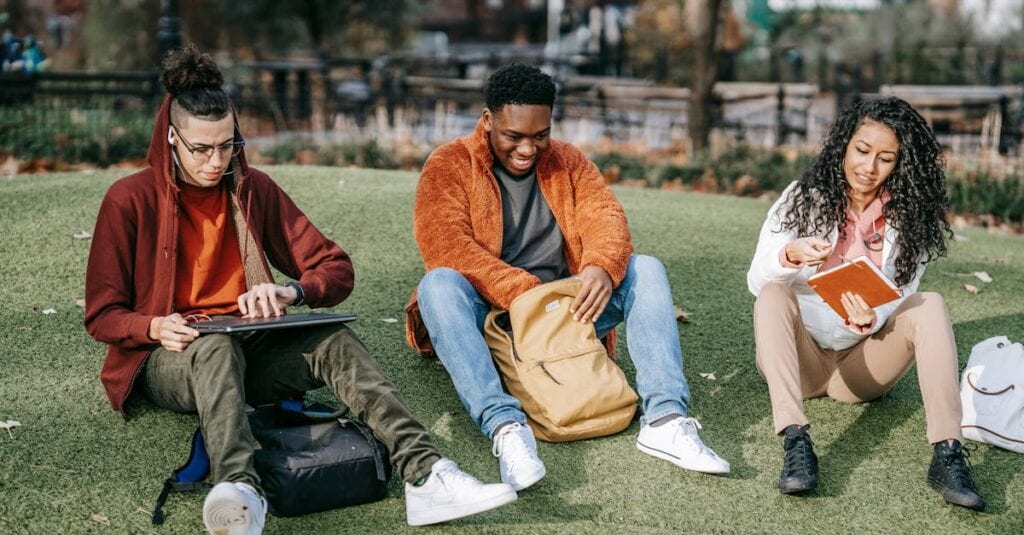 Full body of multiracial classmates in trendy clothes sitting on grass and studying with notepads and laptop