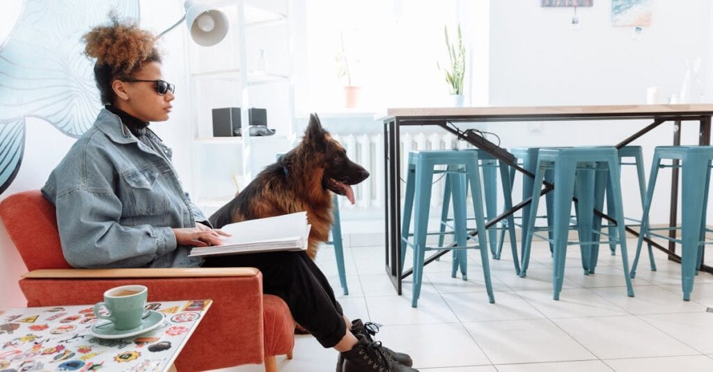 A visually impaired woman sits reading a braille book beside her guide dog indoors.