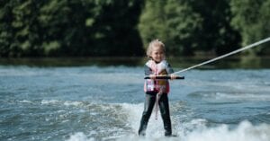 Cheerful young girl wakeboarding on a sunny summer day, sporting a life vest and enjoying water sports on a scenic lake.