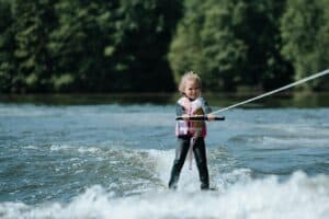 Cheerful young girl wakeboarding on a sunny summer day, sporting a life vest and enjoying water sports on a scenic lake.