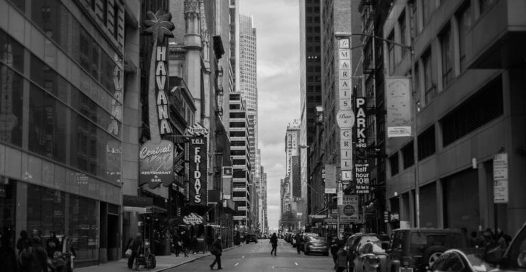 Black and white cityscape of a busy urban street in New York City with skyscrapers and signs.
