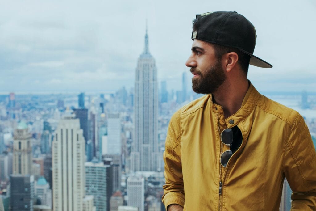 Man with sunglasses and cap stands against New York City's iconic skyline featuring the Empire State Building.