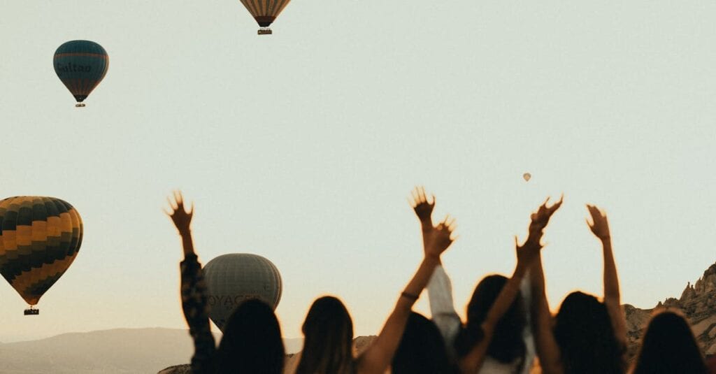 Silhouettes of women watching hot air balloons at sunset, arms raised in excitement.