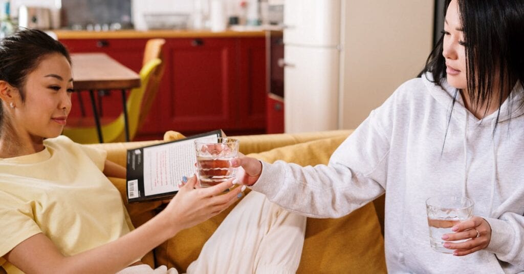 Two friends enjoying a relaxed moment at home, reading and sharing drinks in a cozy living room.