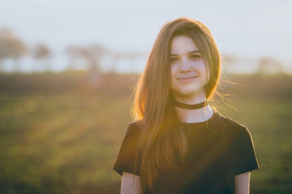 Portrait of a smiling young woman outdoors in a sunlit field, wearing a black choker.