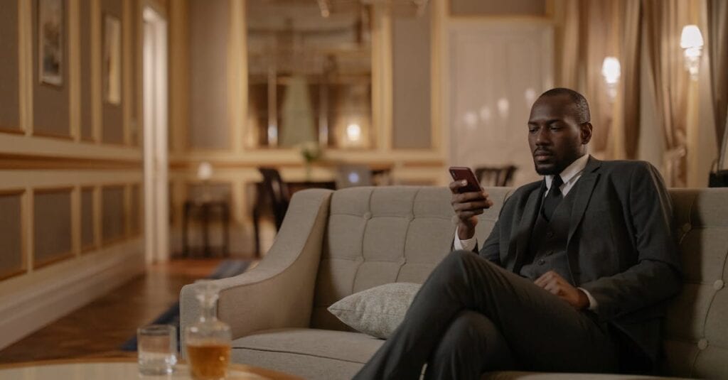 Elegant businessman using smartphone while relaxing in hotel lounge with whiskey on the table.