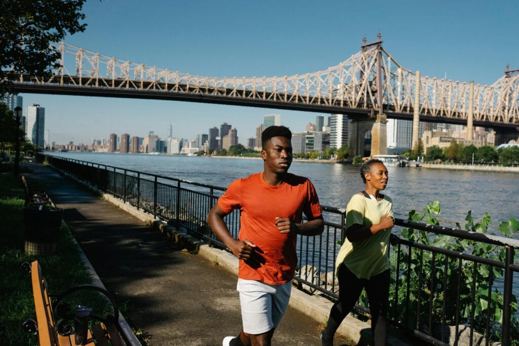 Dynamic shot of a couple jogging by a city river with a prominent bridge in the background.