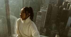 A serene portrait of a woman in a white sweater atop a New York skyscraper with city skyline.
