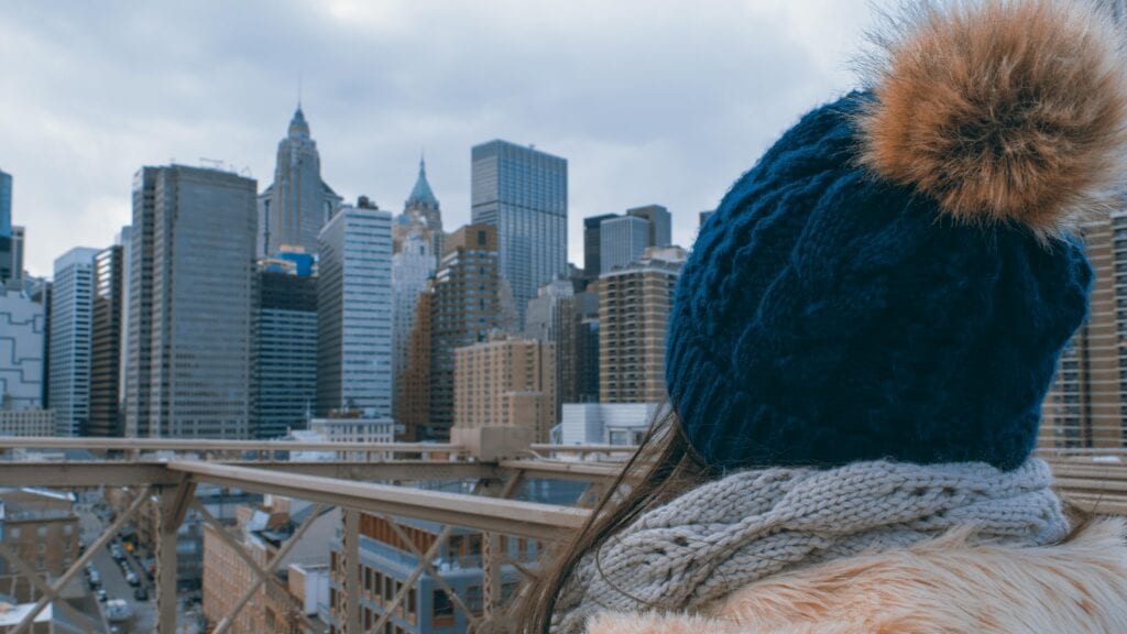 A woman in a winter hat looks at the city skyline from a bridge with clear blue skies.