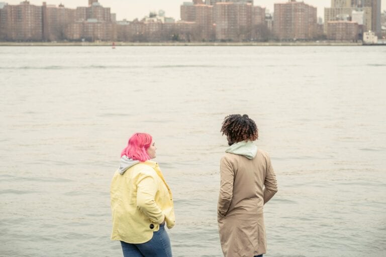 Two friends enjoying a peaceful moment by the riverbank with a cityscape in the background.