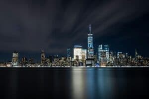 Illuminated New York City skyline featuring One World Trade Center reflecting in the river at night.