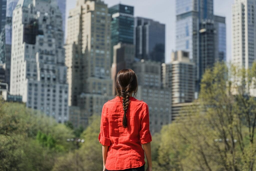 A woman with braided hair in a red top looks at towering city skyscrapers.