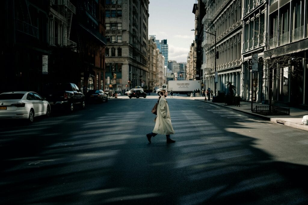 A woman in a trench coat walks across a sunlit street in New York City with tall buildings surrounding her.