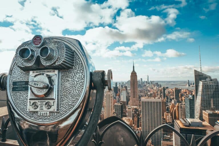 New York Blogs | Articles | News | Tips & Tricks | Video | FAQ | Information | Infograph. 13 View of New York City skyline featuring iconic Empire State Building with observation binoculars in the foreground.