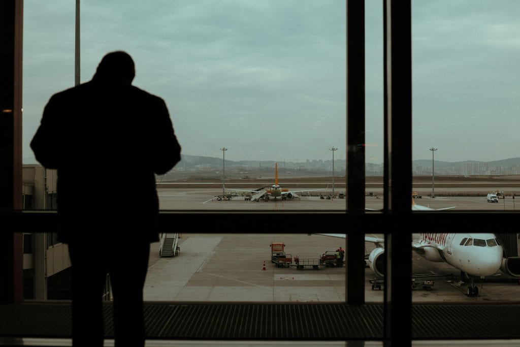 A silhouetted man observes planes and runway through large glass windows at an airport.