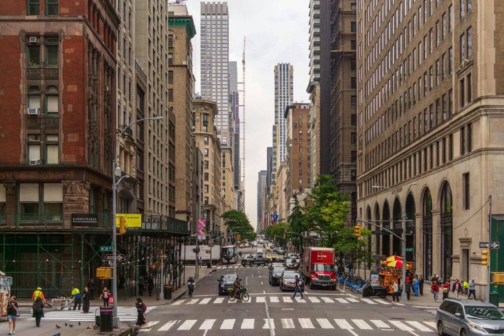 A busy street view of Midtown Manhattan with historic and modern buildings, pedestrians, and traffic.