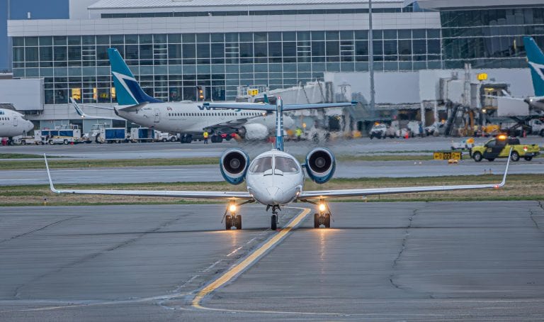A private jet on the runway at a bustling airport with commercial planes and terminal in view.