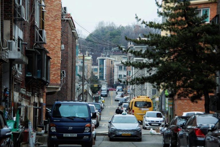 A bustling narrow street lined with parked cars and urban architecture.