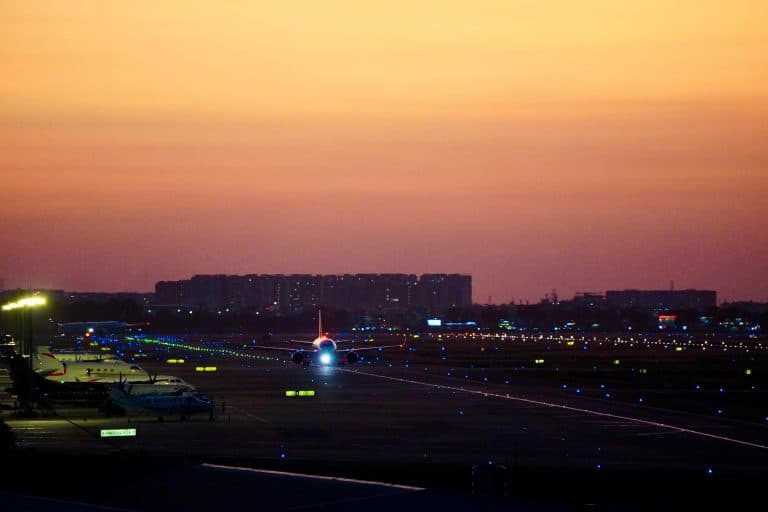 Car Service Blogs | Articles | News | Tips & Tricks | Video | FAQ | Information | Infograph. 17 Airplane preparing for takeoff on an illuminated runway, against a sunset cityscape backdrop.