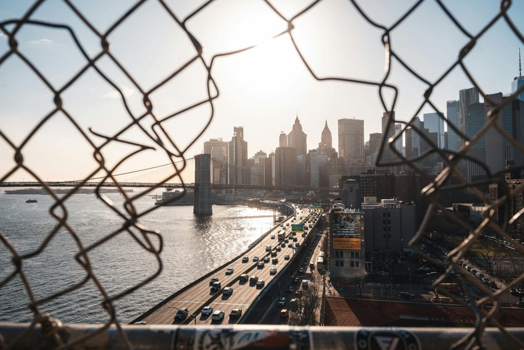 A view of the New York City skyline through a fence with a bridge and roadway below, capturing urban life.