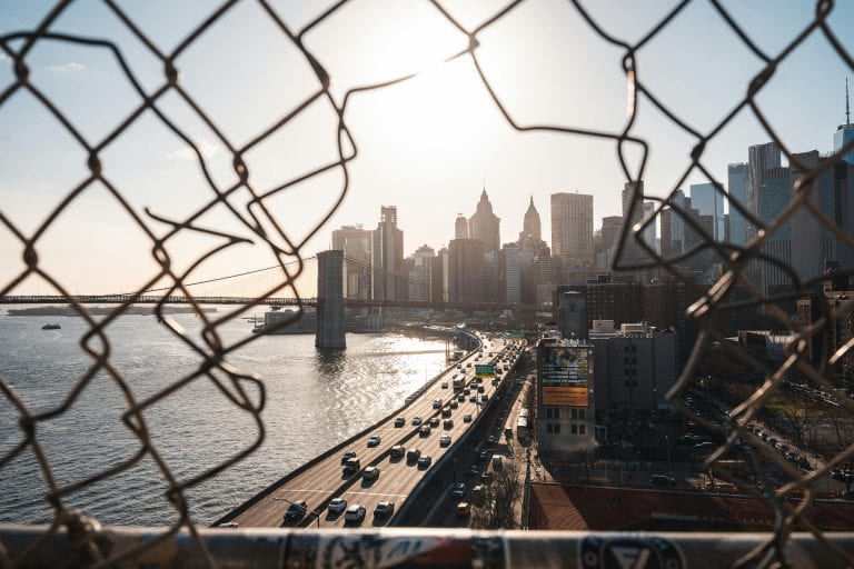 A view of the New York City skyline through a fence with a bridge and roadway below, capturing urban life.