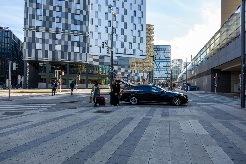 A bustling urban intersection with modern buildings, pedestrians, and a black car in daylight.